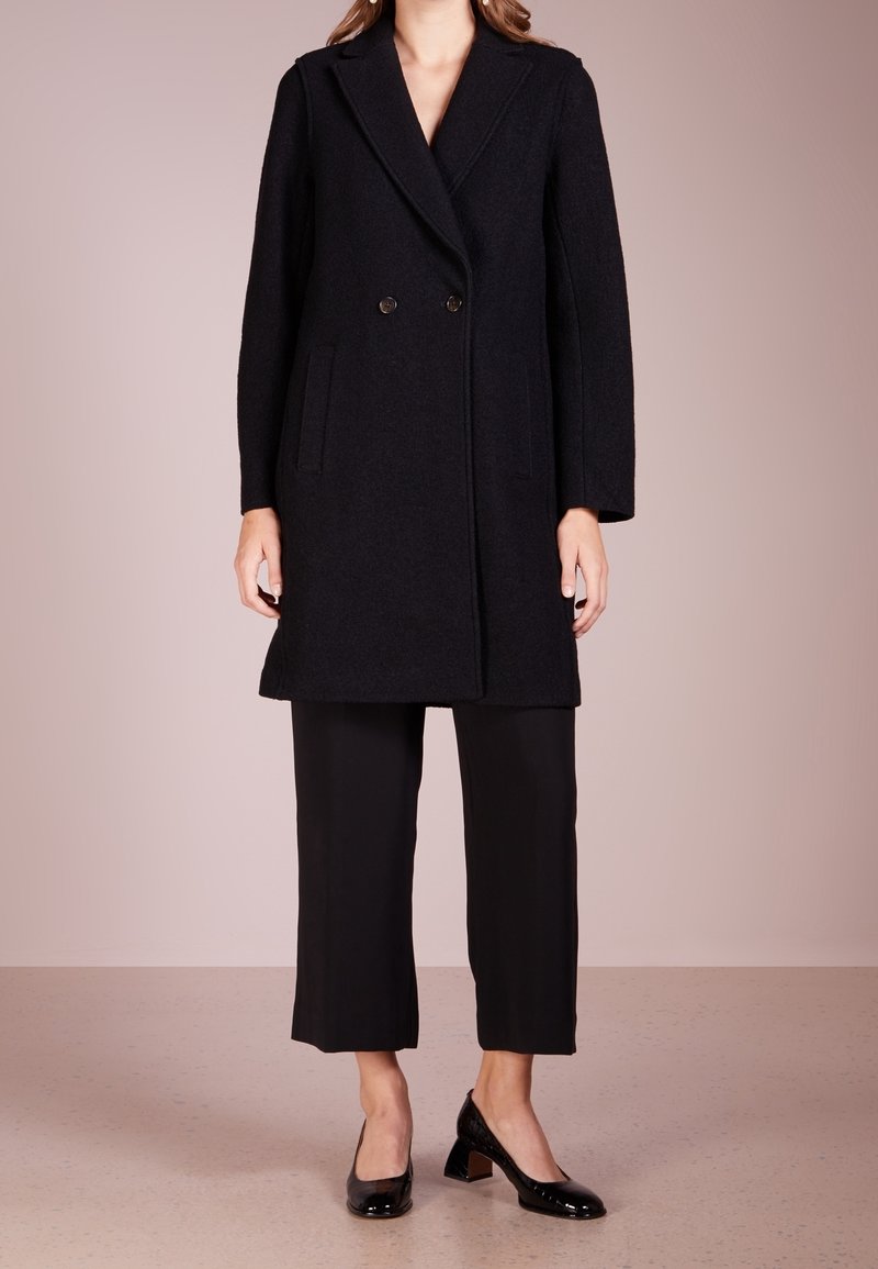 Femme portant un manteau noir à double boutonnage longueur genou, pantalon noir court et chaussures noires vernis à talons bas, debout sur un sol neutre.