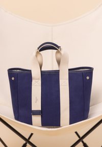 Blå canvas shoppingväska med vita remmar. Har ett handtag upptill, rundade kanter och silverfärgade detaljer. Sytt mönster längs remmarna.