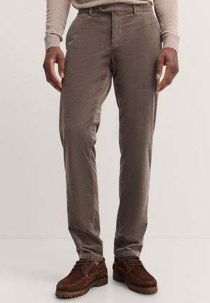Braune Cordhose mit vertikalen Streifen, geradem Schnitt, zwei Fronttaschen und Knopfverschluss. Getragen mit braunen Schnürschuhen.