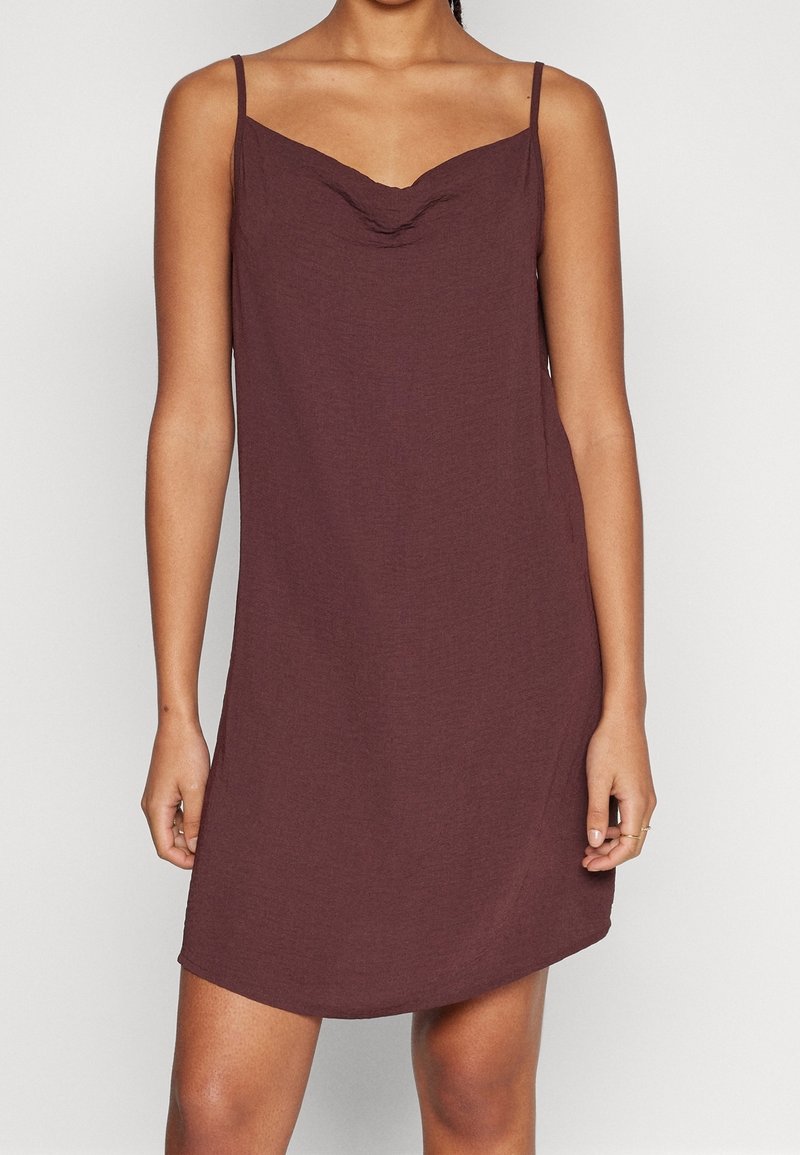Femme portant une robe marron sans manches, longueur genou, avec de fines bretelles et un décolleté subtilement drapé.