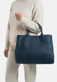 Leren schoudertas in diepblauw; beschikt over twee handvatten aan de bovenkant, twee voorzakken en subtiele gestikte details op het lichaam. Gladde textuur.