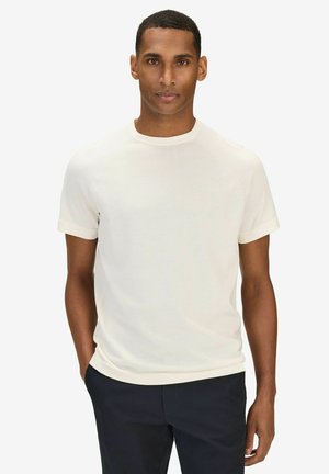 Mann mit kurzen Haaren, der ein eng anliegendes weißes Kurzarmhemd und dunkle Hosen trägt, stehend mit einer Hand in der Hosentasche.