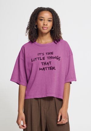 Giovane donna dai capelli ricci che indossa orecchini a cerchio e una maglietta viola con il testo "It's the little things that matter", in piedi davanti a uno sfondo semplice.