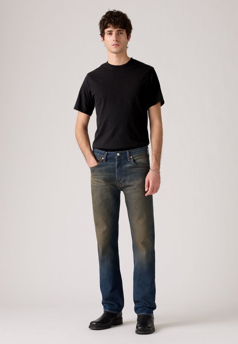 T-shirt noir à manches courtes associé à un jean bleu délavé ; le jean présente une légère usure, deux poches avant et une fermeture à bouton. Bottes noires.