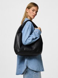 Fekete szövött bőr táska laza kialakítással, texturált mintákkal és széles vállpánttal, egy farmeringes személy által tartva.