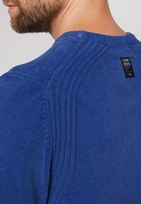 Maglione blu in tessuto strutturato, caratterizzato da un colletto a coste, cuciture dettagliate sulle spalle e un'etichetta nera sul retro.