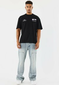 Camiseta negra de manga corta con logotipos gráficos en contraste blanco, combinada con unos pantalones vaqueros de corte holgado en azul claro que presentan bolsillos de parche y una textura sutil.