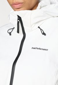 Casaco branco com capuz, fecho de correr preto e cordões. Apresenta um logo preto "PeakPerformance" no peito. Textura suave.