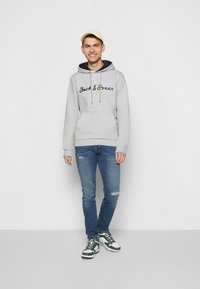 Grå hættejakke med mørkeblå indvendig foring, "Jack & Jones"-logo i marineblå og gul. Båret med blå jeans og grønne og hvide sneakers.
