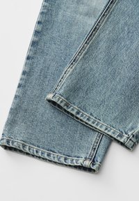 Jean en denim bleu clair, plié en bas, montrant des coutures détaillées et la texture du tissu sur fond blanc.