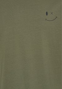 Tela verde olivo con un pequeño diseño de cara sonriente negra, que presenta un ojo ovalado y un ojo tachado, situado cerca de la esquina superior derecha.