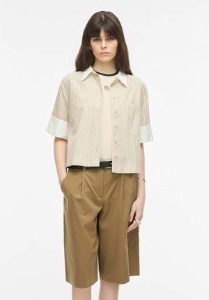 Młoda kobieta z długimi brązowymi włosami, ubrana w beżową koszulę w paski z krótkim rękawem na jasnobeżowym topie, brązowe szorty do kolan i czarny pasek, dłonie w kieszeniach.