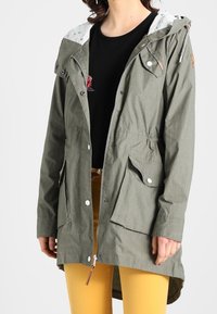 Oliwkowa kurtka z kapturem, z przednimi kieszeniami i zatrzaskami. Posiada wzorzystą podszewkę i ściągnięty w pasie krój. Długi, prosty fason.