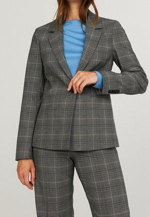 Femme portant un blazer à carreaux gris et un pantalon assorti avec une chemise bleu clair en dessous, ajustant le bouton avant du blazer.