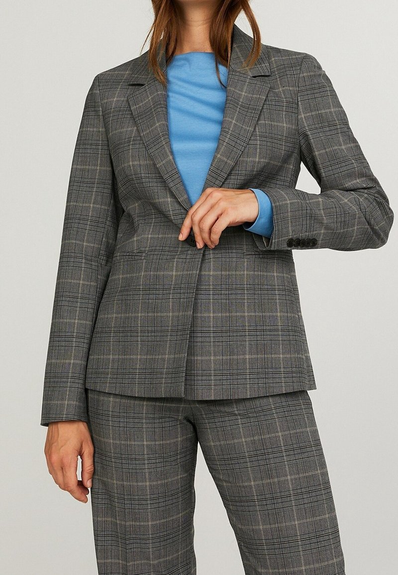 Femme portant un blazer à carreaux gris et un pantalon assorti avec une chemise bleu clair en dessous, ajustant le bouton avant du blazer.