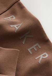 Bruine stof met verhoogde zwarte letters die "BAKER" in een diagonale lay-out spellen, met textuur en stikseldetails aan de rand.