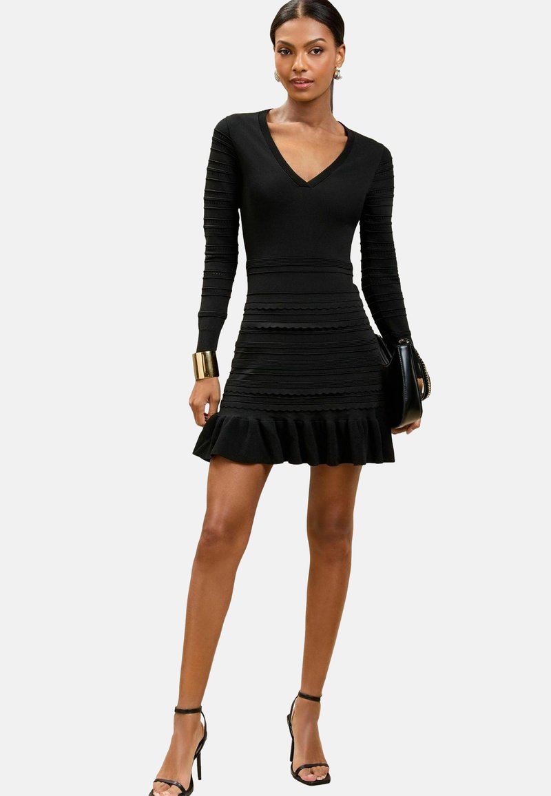 Schwarzes Kleid mit langen Ärmeln, V-Ausschnitt, strukturiertem Rippendesign, tailliertem Schnitt und geschichteter Rüsche am Saum. Kombiniert mit einer schwarzen Handtasche und High Heels.
