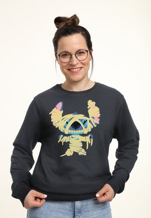Kvinde med briller og håret samlet i en knold iført en mørk sweatshirt med en farverig tegneseriefigur af en mumie trykt på fronten.