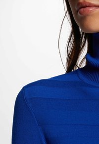 Hochgeschlossenes Sweatshirt in leuchtendem Blau mit horizontaler Rippstruktur. Eine Nahaufnahme hebt das Design des Kragens und die glatte Stoffdetail hervor.