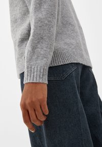 Grå stickad tröja med ribbade ärmar, kombinerad med mörkblå jeans. Fokus på ärmarnas textur och tygdetaljer.