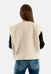 Maglione senza maniche beige con una finitura in lana texturizzata, abbinato a jeans blu aderenti con un design cucito distintivo sulle tasche posteriori.