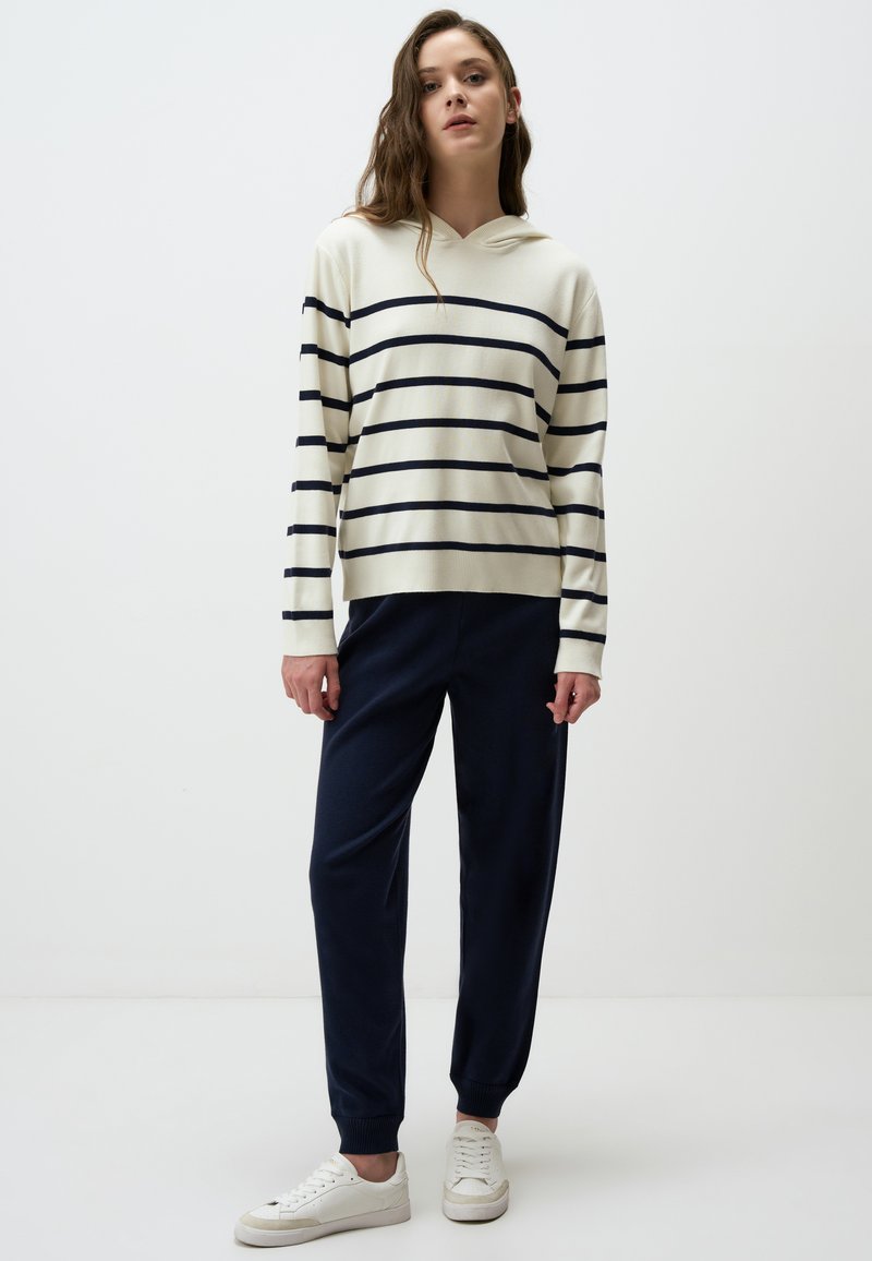 Sweatshirt à capuche crème avec des rayures marine, associé à un pantalon tapered marine et des baskets blanches. Texture lisse, coupe décontractée et poignets élastiques.