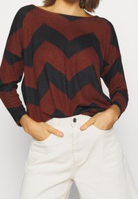 Langarmshirt mit einem rot-schwarz gestreiften Zickzack-Muster, aus weichem Stoff gefertigt. Kombiniert mit hoch taillierten weißen Hosen mit Knopfdetails.