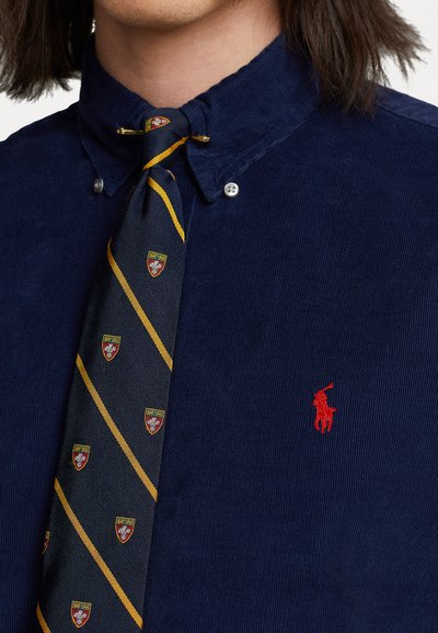 Marineblå kordfløyelskjorte med knapp-down krage; har liten rød polo-logo. Knyttet med et mønstrete slips i marineblått, gult og heraldisk design.