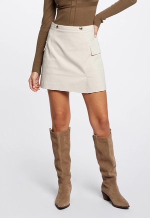 Vrouw draagt een beige minirok met zijzak en beige kniehoge suède laarzen, gecombineerd met een bruin geribd topje met lange mouwen.