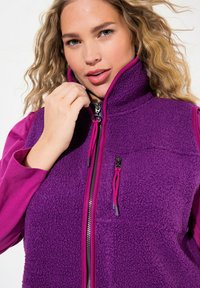 Lila Fleece-Weste mit hohem Kragen, Reißverschluss vorne und pinken Akzenten. Mit strukturiertem Stoff und Kordelzug am Halsausschnitt.