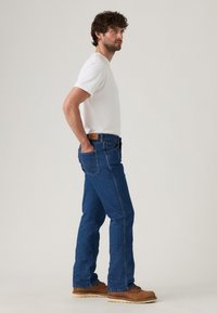 Jeans de denim azul con corte recto, que presentan una parche de cuero color beige en la cintura trasera y bolsillos laterales. Llevados con una camiseta blanca.