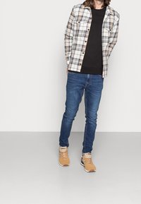Rutig flanellskjorta i beige, grått och marinblått; svart T-shirt med rund hals under; blå jeans; tan sportskor med vita detaljer.
