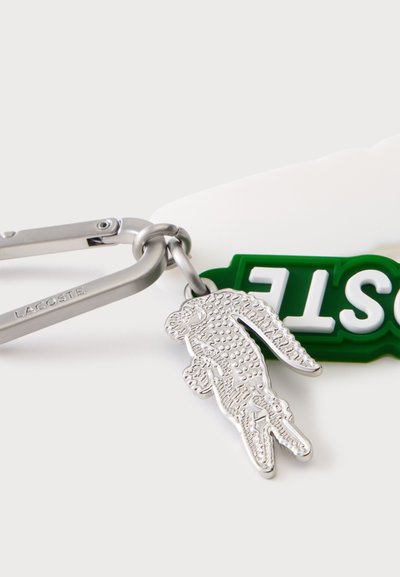 Сребърен карабинер с марка Lacoste държи детайлна сребърна висулка крокодил и зелен етикет с бели изпъкнали букви.