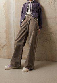 Giacca in pile viola con zip, pantaloni cargo beige e scarpe platform bianche. Texture liscia, vestibilità comoda e tasche funzionali.
