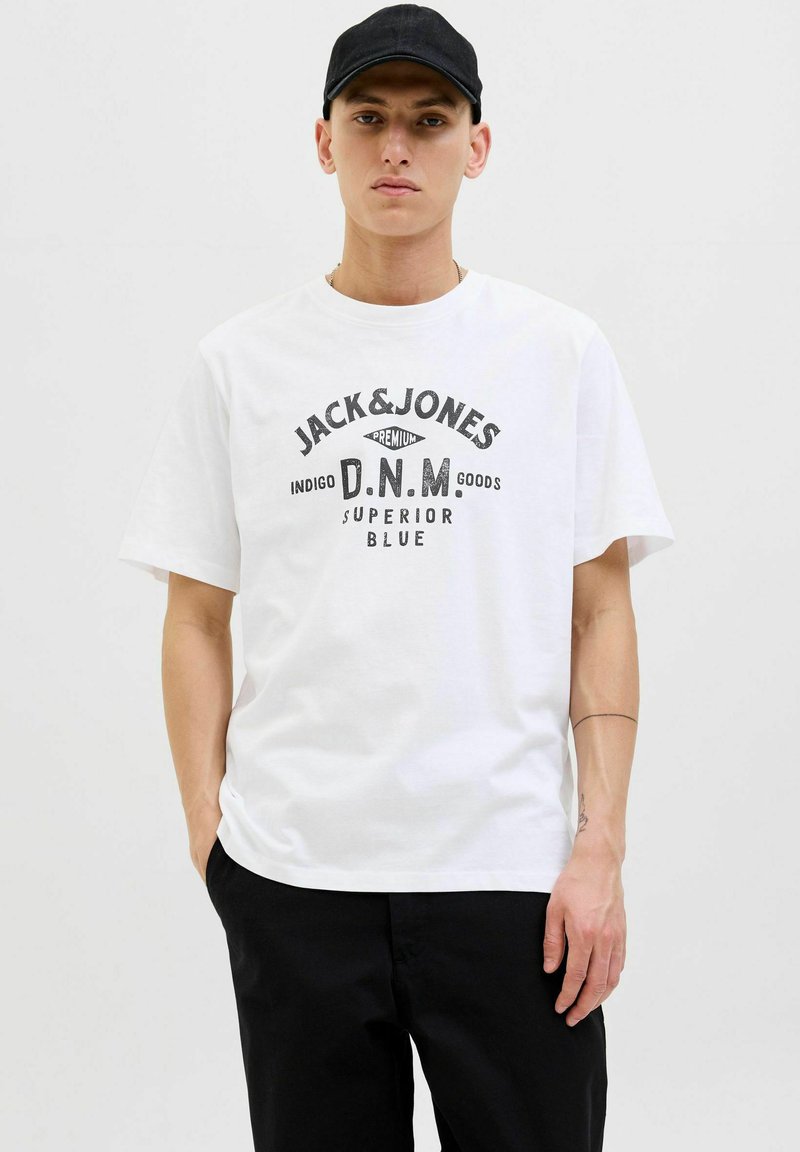 Balta medvilninė marškinė su trumpomis rankovėmis, ant kurių spaudintas juodas logotipas su užrašu "JACK & JONES D.N.M. GOODS SUPERIOR BLUE." Dėvima su juodomis kelnėmis.