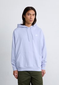 A person with long hair wears a light purple hoodie with a small white Nike logo, standing against a plain background.