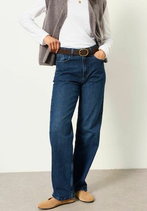 Frau trägt blaue Jeans, weißes Langarmshirt, graue Weste, braunen Gürtel und beige Slip-On-Schuhe, steht mit Händen in den Taschen.