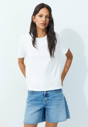 Kvinde med langt mørkt hår iført en simpel hvid t-shirt og blå denimshorts, stående mod en hvid baggrund.