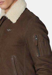 Bruine leren jas met een witte schapenvacht kraag, een ritszak op de mouw en een metalen embleem op de borst. Soepele textuur.