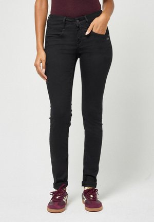 Woman wearing high-waisted black skinny jeans with rolled cuffs and burgundy sneakers, standing against a plain light background.