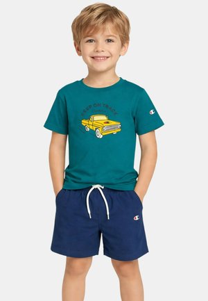 Ragazzo sorridente che indossa una maglietta turchese con grafica di un camion giallo e pantaloncini blu scuro con coulisse bianca, mani nelle tasche.