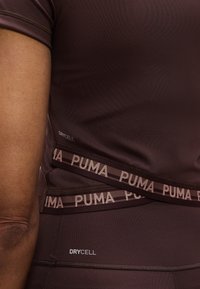 Κοντινό πλάνο ενός ατόμου που φοράει σκούρα καφέ αθλητική ενδυμασία Puma με το κείμενο "DRYCELL" και ελαστικές ταινίες με το λογότυπο γύρ ω από τη μέση.