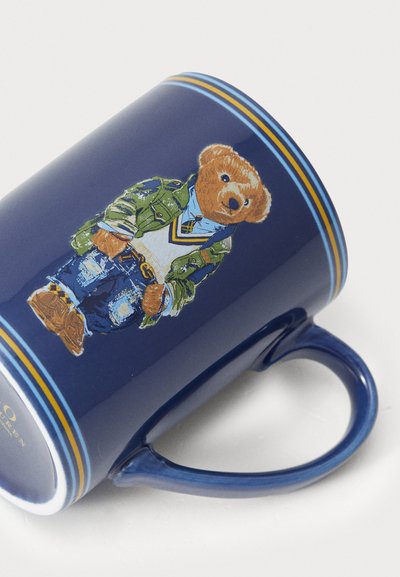 Tasse en céramique navy avec une finition brillante, présentant un graphique d'un ours en veste verte et en jeans bleus, avec des accents de rayures jaunes.