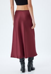 Gonna maxi in satin bordeaux con una texture liscia, silhouette svasata e una vita semplice, abbinata a stivali neri fino al ginocchio.