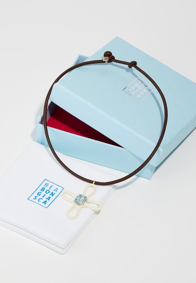 Collana in corda marrone con un pendant a forma di fiore bianco e centro in pietra blu. Sistemata con una scatola azzurro chiaro e un panno bianco.