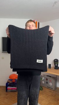 Svart strikket skjerf med en ribbestruktur, med en fremtredende etikett som leser "DRYKORN" nederst til høyre.