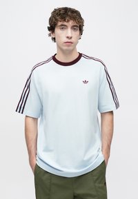 Ανοιχτό μπλε μπλουζάκι με λαιμόκοψη λαιμού, μωβ μπορντό ρίγες στα μανίκια και κεντημένο λογότυπο στο στήθος. Κατασκευασμένο από μαλακό, υφασμάτινο ύφασμα με υφή.