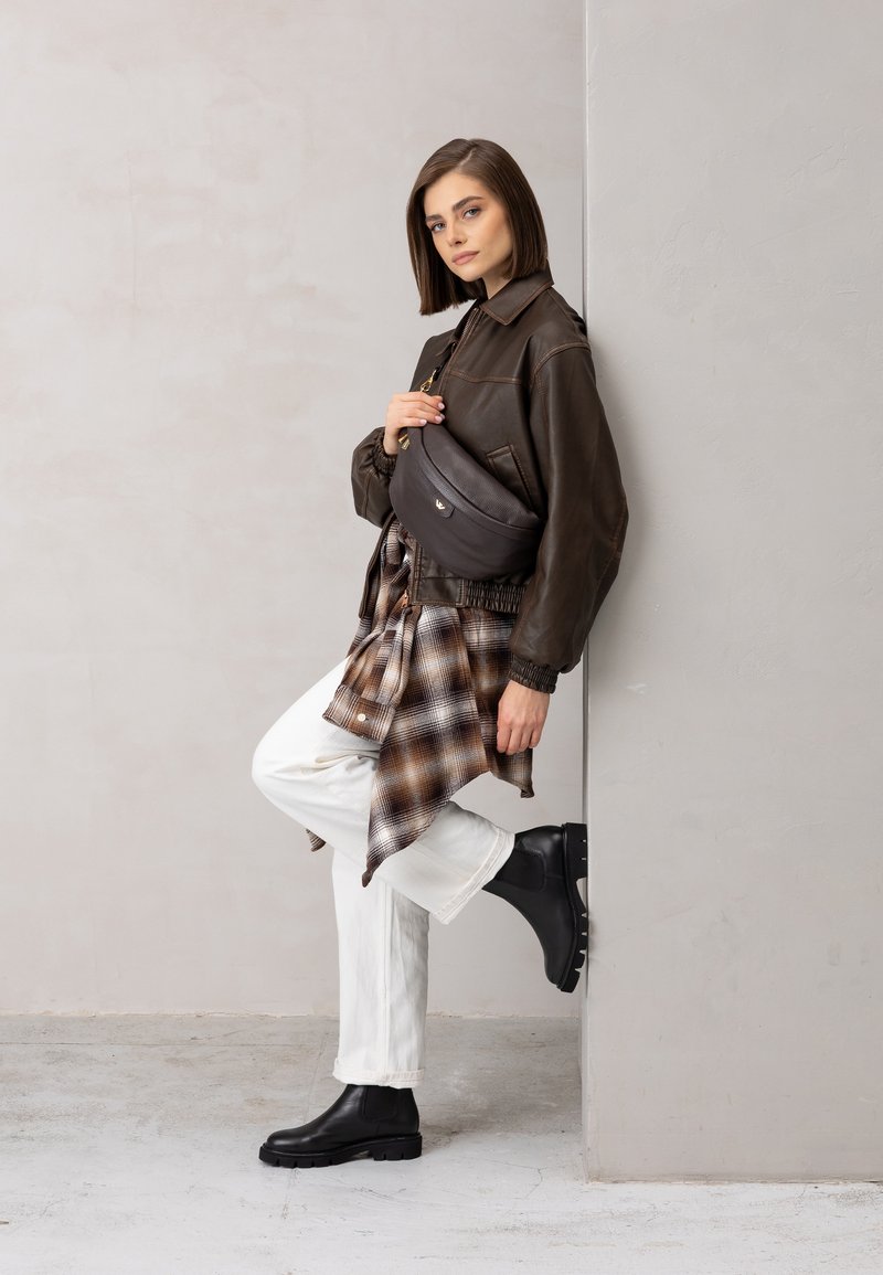 Bruin leren jack, witte loszittende broek, zwarte enkellaarsjes en een bruine crossbodytas. De outfit wordt compleet gemaakt met een geruit overhemd dat om de taille is geknoopt.