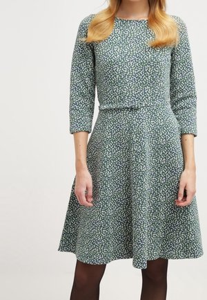 Kvinde iført en knælængde, grøn mønstret kjole med trekvart-ærmer og sorte strømpebukser, stående mod en ensfarvet baggrund.