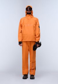 Person i orange snedragt, bukser, sorte støvler og skibriller, der holder sorte handsker og står mod en ensfarvet lys baggrund.